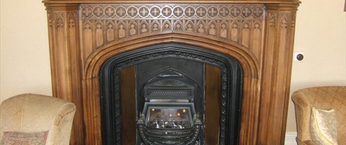 Fireplace with a carved wooden surround.  The arched opening is a four-centred arch with spandrels above containing a series of a repeated motif, representing the tall, thin arched window of a Gothic church.